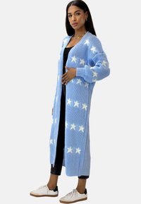 Cárdigan de punto de color azul claro con patrones de estrellas blancas, mangas largas y diseño de frente abierto, combinado con leggings y zapatos negros.