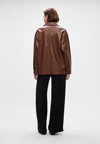 Jaqueta de couro castanho com um corte descontraído, colarinho grande e punhos abotoados, combinada com calças largas pretas, vista pela parte de trás.