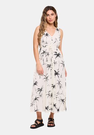 Mujer con vestido largo blanco sin mangas con estampados de palmeras negras y sandalias negras de punta abierta, de pie contra un fondo claro y liso.