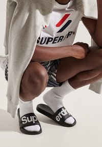 Černé skluzavkové sandály s bílým textem "Super", nošené s bílými ponožkami. Oblečení zahrnuje šedou mikinu a vzorované kraťasy.