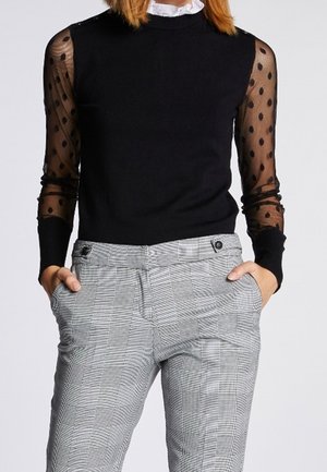 Frau trägt einen schwarzen Pullover mit transparenten, gepunkteten Ärmeln und eine hoch taillierte graue karierte Hose, die Hände in den Taschen.