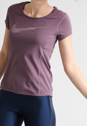 Kvinde iført en lilla kortærmet Nike sportstrøje og marineblå træningsleggings med sidelommer mod en ensfarvet baggrund.