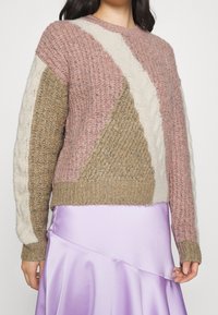 Strickpullover mit geometrischem Design in Pink, Creme und Beige. Die Textur umfasst Zopfmuster-Details und einen gerippten Saum.