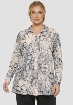 Femme portant un chemisier à manches longues boutonné avec un motif serpent noir et beige, debout devant un fond gris clair uni.