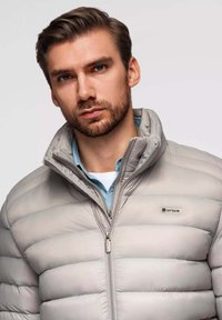 Hellgraue Pufferjacke mit horizontalen Nähten, Stehkragen und Frontreißverschluss. Getragen über einem hellblauen Hemd. Logo auf der Brust.