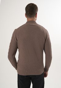 Man in een getextureerde, bruine gebreide trui met geribbelde kraag en boorden, gezien vanaf de achterkant tegen een effen witte achtergrond.