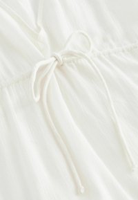Witte, lichte stof met een gestructureerde afwerking, voorzien van een gestrikte trekkoorddetail bij de taille en subtiele plooien langs de naad.
