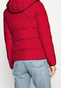 Rote Steppjacke mit Kapuze, mit einem gesteppte Design und elastischen Bündchen. Kombiniert mit hellblauen Jeans.