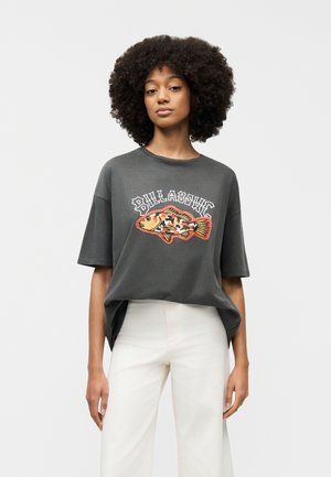 Femme aux cheveux bouclés portant un t-shirt ample gris foncé avec un motif de poisson coloré et un pantalon blanc, debout devant un fond uni.
