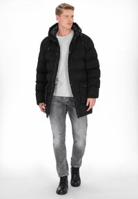 Czarna puchowa kurtka z kapturem, zapinana na zamek błyskawiczny, z przednimi kieszeniami. Nosi się ją na szarym swetrze, w połączeniu z szarymi dżinsami i czarnymi butami.