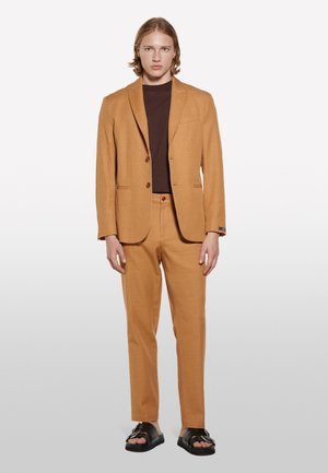 Un costume beige à coupe slim, avec une veste à deux boutons et un pantalon assorti, associé à un haut marron et des sandales noires. Texture lisse, design sur mesure.