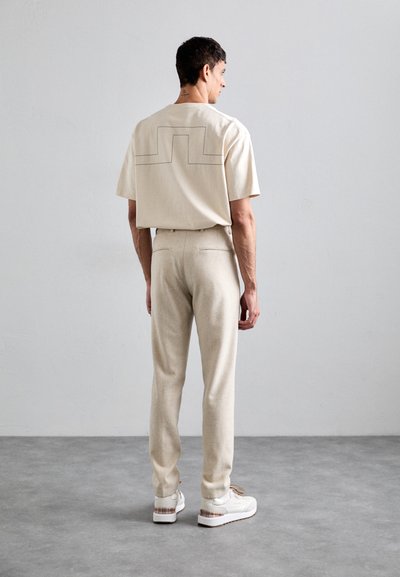 Man standing indoors facing away, wearing beige pants, a beige short-sleeve shirt with a black line design on the back, and white sneakers.