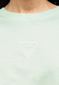 Κοντινό πλάνο λαιμού μπλουζάκι σε ανοιχτό πράσινο χρώμα με ανάγλυφο υφασμάτινο τρίγωνο λογότυπο, στο κέντρο κάτω από την κλείδα, με την επιγραφή "GUESS".