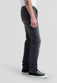 Person stående iført mørkegrå slim-fit jeans, sorte og hvide lærreds-sneakers og en hvid skjorte, med en tatovering på underarmen.