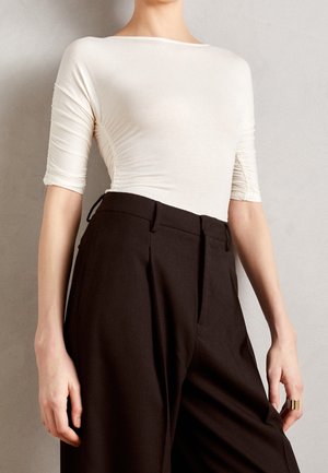 Kobieta w dopasowanym, kremowobiałym topie z rękawami do łokci i wysokim stanem oraz szerokich, ciemnobrązowych spodniach, stojąca na tle neutralnej ściany.