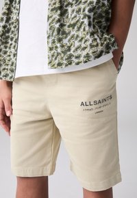 Beige bomullsshorts med sydd fåll, med "ALLSAINTS LONDON"-tryck i mörkt på vänster sida. Bärs med en mönstrad grön skjorta.