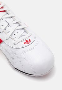 Adidași din piele albă cu accente roșii, având vârful rotund, un design stratificat și un logo distinctiv lângă șireturi. Suprafață texturată.