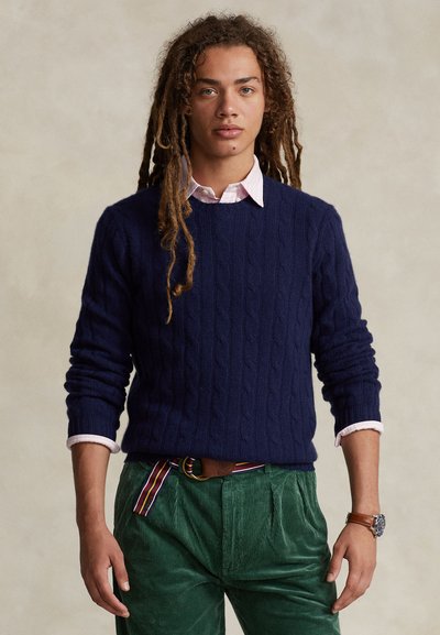 Μπλε πουλόβερ με πλέξη καλώδιο και στρογγυλή λαιμόκοψη, φορεμένο πάνω από ένα ανοιχτό ροζ πουκάμισο με γιακά. Πράσινο παντελόνι κοτλέ και καφέ ζώνη με λεπτομέρειες.