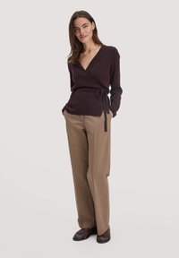 Burgunderfarbener Wickelpullover mit langen Ärmeln und seitlichem Knoten; kombiniert mit beigen geraden Hosen und braunen Schuhen. Glatte Textur, lässige Passform.