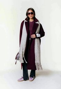 Casaco bordeaux com colarinho notched, cachecol cinza com franjas, jeans escuros e ténis lilás; modelo a usar óculos de sol oversized.