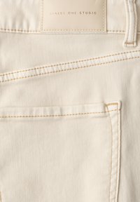 Poche arrière d’un jean en denim écru avec des surpiqûres orange visibles et une étiquette beige cousue portant la mention "STREET ONE STUDIO."