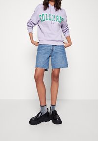 Persona che indossa un felpa lavanda con la scritta "COLORADO" in verde, pantaloni shorts di jeans con orli sfrangiati, calze argento e mocassini neri a piattaforma chunky.
