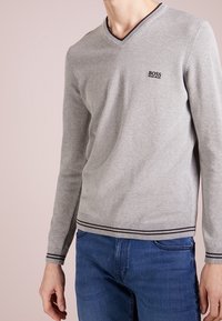 Grauer V-Ausschnitt-Pullover aus weichem Strickmaterial mit einem Logo auf der Brust, versehen mit schwarzen Streifenakzenten am Halsauschnitt und Saum.