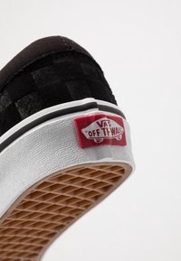 Talons de chaussure Vans noirs montrant la semelle blanche texturée et l'étiquette rouge avec le logo « Vans Off The Wall », vue de dessous sur fond blanc.