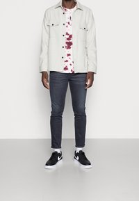 Camisa de botones gris claro con dos bolsillos en el pecho sobre una camiseta de tie-dye blanca y granate, combinada con jeans oscuros y zapatillas negras.