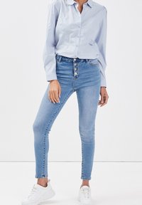 Jean taille haute, bleu clair, coupe ajustée, fermeture avant à cinq boutons et légère décoloration, associé à une chemise bleu clair et des baskets blanches.