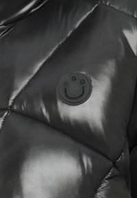 Čierna puffa bunda s lesklým povrchom, diamantovým prešívaním a okrúhlym čiernym logom s usmievajúcou sa tvárou na hrudi.