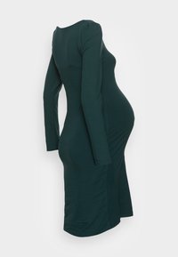 En figursydd mörkgrön långärmad graviddress som visas på en huvudlös mannekäng mot en enfärgad bakgrund.