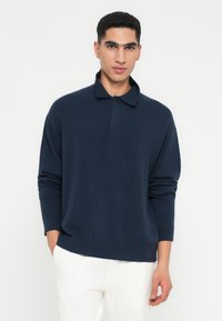 Giovane con capelli neri ricci che indossa una polo a maniche lunghe blu navy e pantaloni bianchi, in piedi con una mano in tasca.