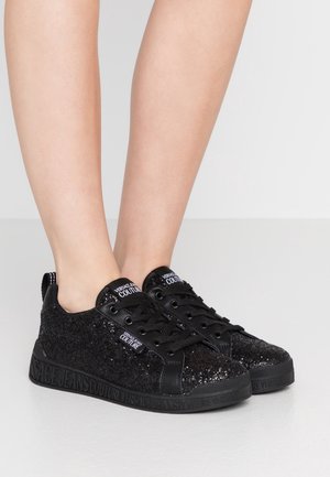Sorte glitrende lavtop-sneakers med sorte snørebånd, "Versace Jeans Couture" mærkater på tungen, siden og præget på sålen, båret på fødder.