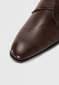 Zapato de vestir marrón de cuero con un acabado suave y un acento de cuero texturizado. Presenta una punta afilada y una suela de goma negra.