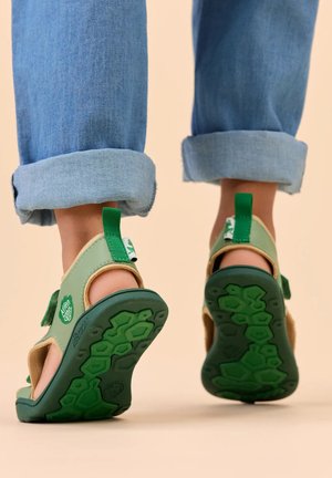 Kind trägt grün-beige offene Sandalen mit strukturierten Sohlen und hochgekrempelte blaue Jeans, läuft auf einer beigen Oberfläche.