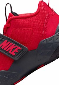 Czerwona tenisówka z teksturowaną cholewką, czarnymi akcentami i wyraźnym czarnym paskiem z logo Nike w kolorze czerwonym. Gruba gumowa podeszwa z geometrycznym wzorem.