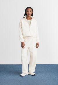 Cream fleece zip-up jacket with high collar, paired with matching cream wide-leg pants. Features casual design and subtle logo detail.