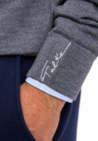 Polsino di una camicia grigia a maniche lunghe con fodera azzurro chiaro, caratterizzato dal testo ricamato "Falke". Il tessuto mostra un motivo a maglia strutturato.