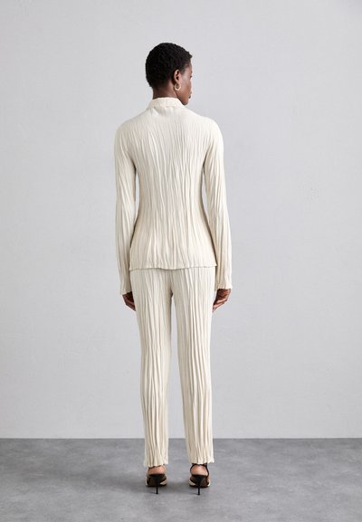 Cream-colored, textured outfit with a fitted top and tapered pants. Features vertical pleats and long sleeves, paired with black heeled sandals.