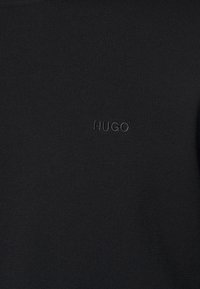 Schwarzer strukturierter Stoff mit dezent dunkel gesticktem "HUGO"-Logo, das zentral auf der Oberfläche platziert ist.