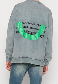 Osoba ubrana w wyblakły niebieski sweter z postrzępionym dołem oraz jasną zieloną abstrakcyjną grafiką i czarnym tekstem na plecach, w połączeniu z niebieskimi dżinsami.