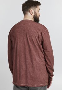 Camicia a maniche lunghe in tessuto rosso scuro con finitura testurizzata. Presenta un collo rotondo e un orlo dritto, con un'etichetta sul lato inferiore.