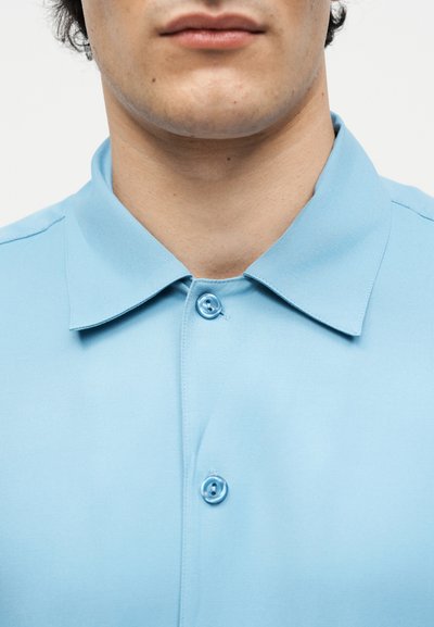 Κοντινό πλάνο στο κάτω μέρος του προσώπου και το λαιμό ενός ατόμου που φορά ένα ανοιχτό μπλε πουκάμισο με κουμπιά και κλασικό γιακά.