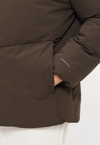 Mørkebrun pufferjakke med tekstureret stof, der har en sidelomme og et logo-label på ærmet. Glat, isoleret design.