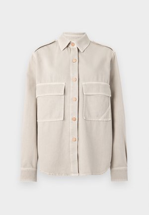 Chaqueta camisa beige con botones y dos bolsillos frontales, usada sobre una camiseta a rayas, combinada con pantalones negros de pierna ancha y bailarinas blancas.