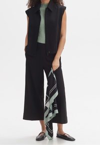 Czarna kamizelka bez rękawów na tle teksturowanej zielonej bluzki, w zestawieniu z szerokimi czarnymi spodniami. Wzorzysty miętowy szalik zwisa z ręki. Czarne baleriny.