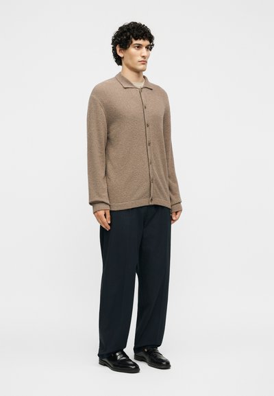 Νέος άνδρας με σγουρά μαλλιά που φοράει καφέ πλεκτή ζακέτα με κουμπιά, μαύρο φαρδύ παντελόνι και μαύρα δερμάτινα παπούτσια, όρθιος ελαφρώς πλαγιαστά.