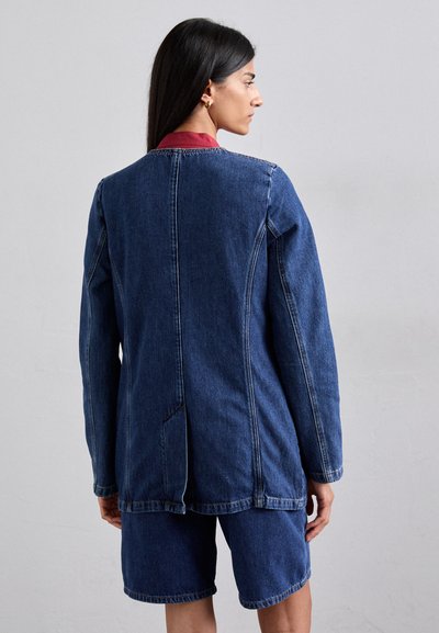 Jachetă din denim cu mâneci lungi, de culoare albastru închis, cu accente roșii la guler și cusături vizibile. Pură peste pantaloni scurți asortați din denim.