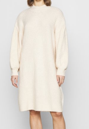 Femme portant une robe-pull en tricot ample, à manches longues, de couleur crème, avec des poignets et un ourlet côtelés, debout devant un fond uni.